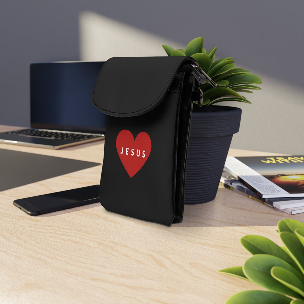 Black Jesus cell phone wallet with red heart design on desk next to plant and laptop