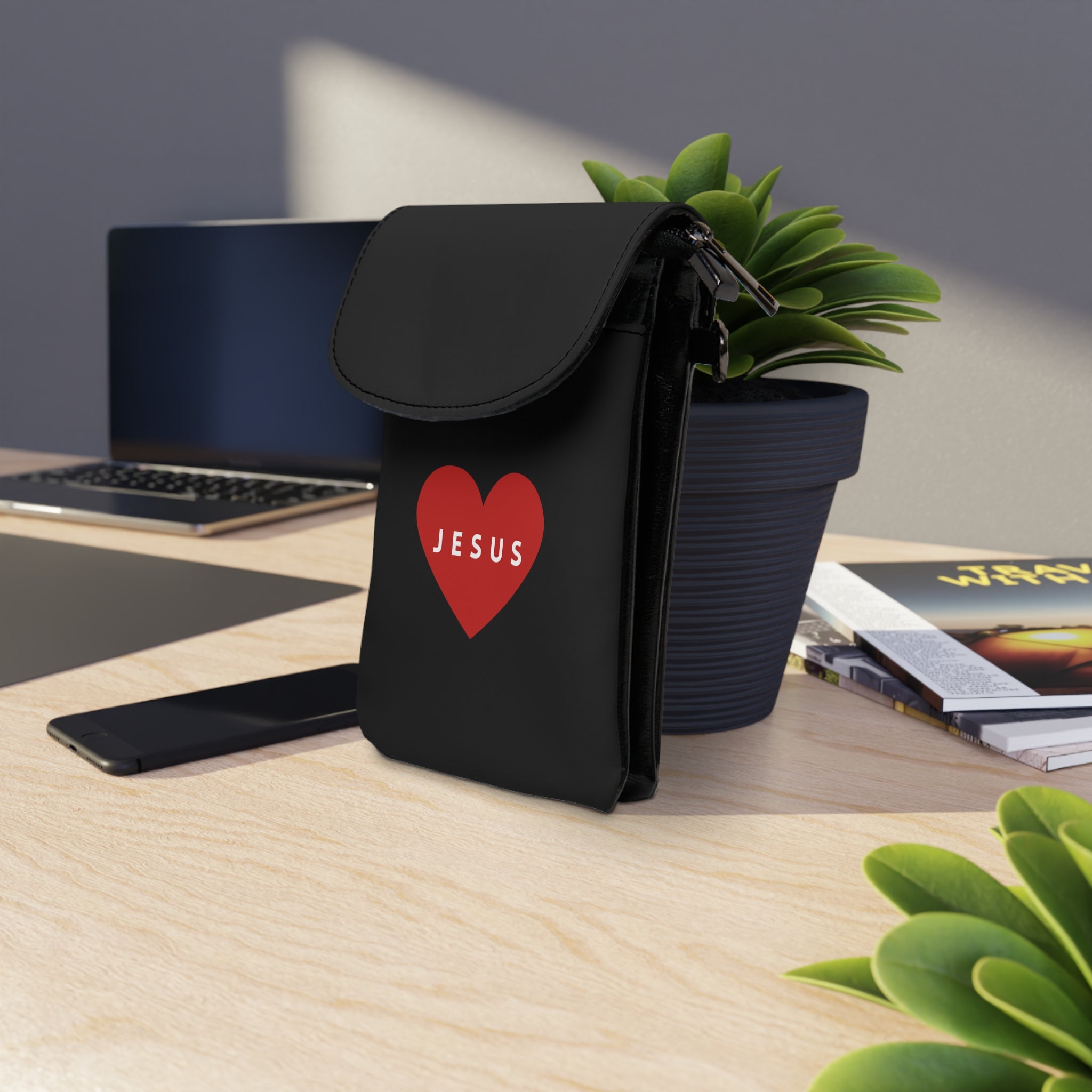 Black Jesus cell phone wallet with red heart design on desk next to plant and laptop