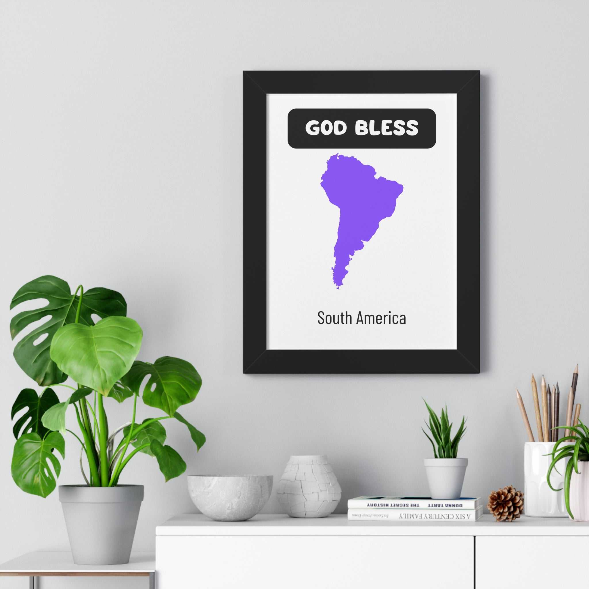 Framed God Bless South America poster with purple map and modern black frame on grey wall above white sideboard with plants and decor