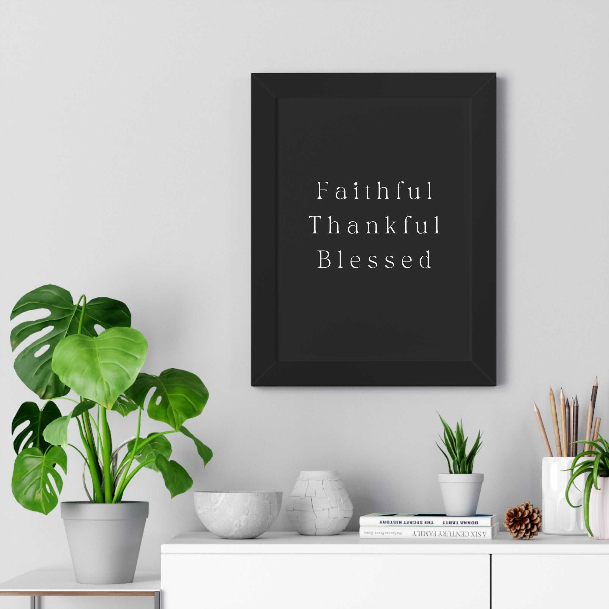 Black framed poster with the words Faithful Thankful Blessed in white text on black background hanging above a white shelf with plants and decor