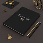 Gratidão Journal black matte cover with gold and black pencils and gold clips on brown textured surface