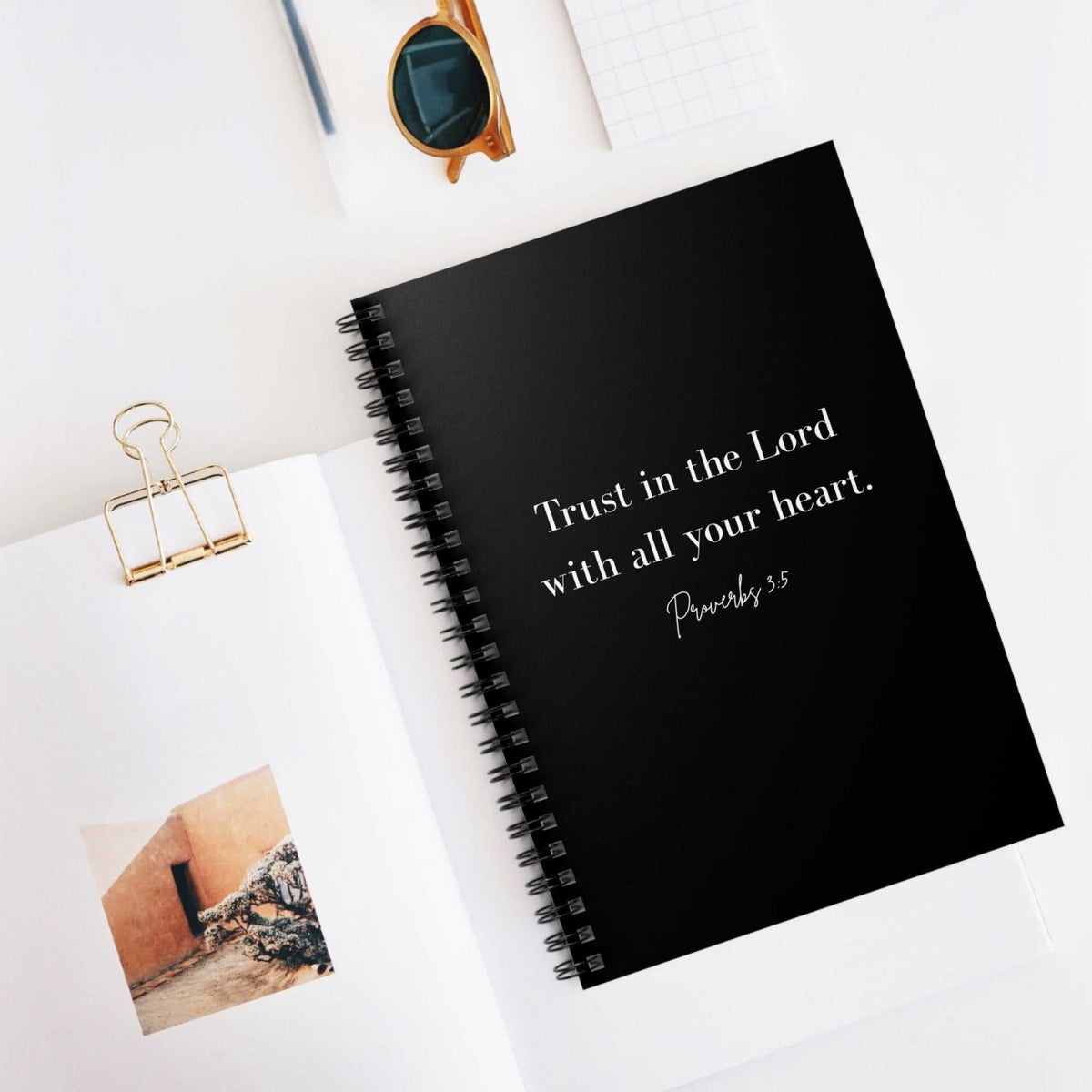 Black spiral bound notebook with "Trust in the Lord with all your heart Proverbs 3:5" printed on the cover, placed on a white surface next to open book and sunglasses